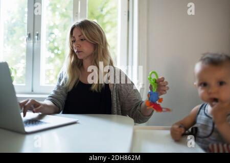 Mère d'un adulte de taille moyenne utilisant un ordinateur portable tout en prenant soin de bébé à la table à manger Banque D'Images