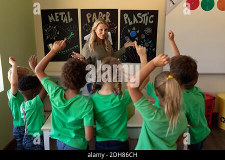 Professeur d'école féminine pointant vers une affiche de recyclage avec un groupe d'écoliers qui élèvent leur ha Banque D'Images