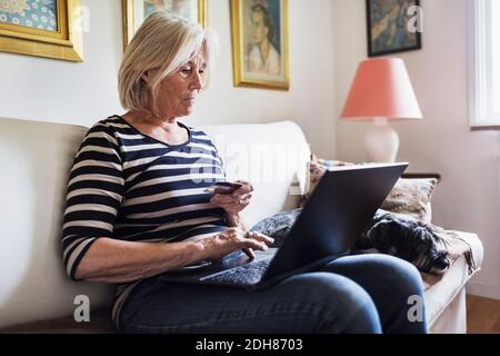 Femme senior magasiner en ligne pendant que le chien se détendant sur le canapé à accueil Banque D'Images