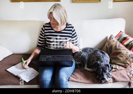 Femme âgée écrivant dans le livre tout en utilisant l'ordinateur portable et le crédit carte par chien sur le canapé à la maison Banque D'Images