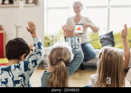 Vue arrière des élèves levant les mains pendant que le professeur affiche le numérique tablette à l'école maternelle Banque D'Images