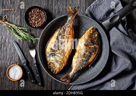 dorade grillée, orata, dorado poisson dans une poêle sur une table en bois sombre avec une fourchette, romarin et poivre, vue horizontale d'en haut, plat Banque D'Images