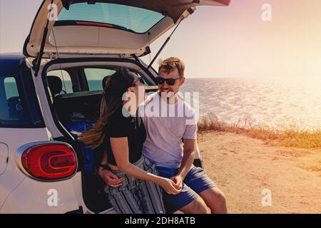 Joyeux jeune couple assis sur le coffre de la voiture près de la mer. Voyage d'été, voyage en voiture Banque D'Images