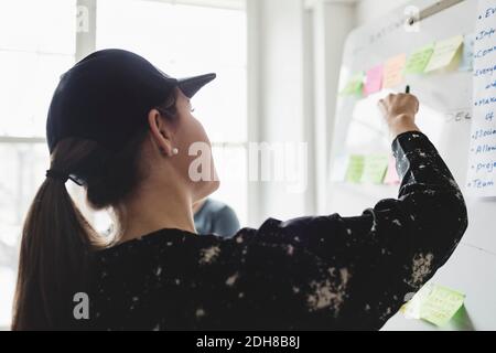 Vue arrière de la femme vérifiant les notes adhésives sur le tableau blanc dans bureau créatif Banque D'Images