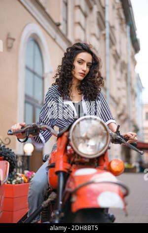 Jeune femme brune en chemise rayée et jeans assis sur une moto rouge à l'extérieur. Banque D'Images