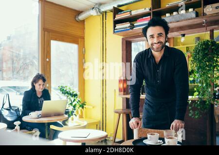 Portrait d'un serveur heureux avec un client utilisant un ordinateur portable en arrière-plan au café Banque D'Images