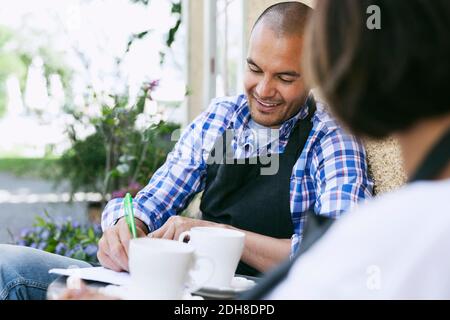 Homme souriant écrivant sur le papier tout en prenant un café avec un collègue café extérieur Banque D'Images