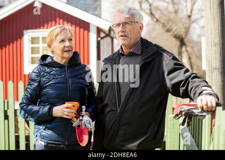 Couple senior souriant tenant l'équipement de jardinage tout en se tenant contre la clôture en hiver Banque D'Images