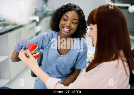 Gros plan horizontal de la jolie jeune femme africaine souriante dentiste, expliquant des façons de traitement ou l'anatomie des dents pour sa patiente, montrant Banque D'Images