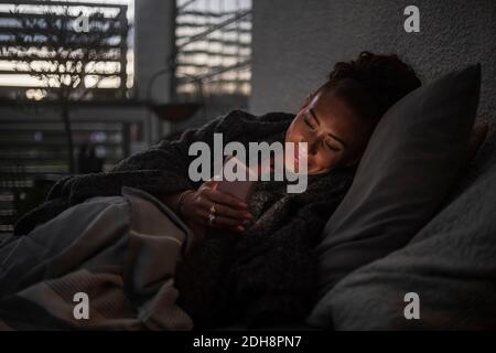 Femme souriante utilisant un téléphone portable sur un canapé à la maison pendant coucher de soleil Banque D'Images