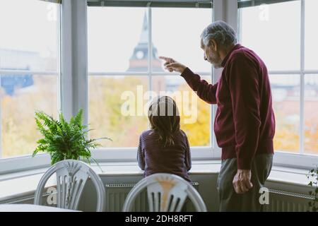 Vue latérale d'un homme âgé montrant quelque chose au grand petit-fils par la fenêtre à la maison Banque D'Images
