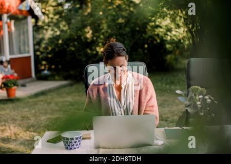 Femme souriante utilisant un ordinateur portable tout en étant assise à la table à manger jardin en été Banque D'Images