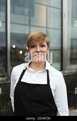 Portrait d'un professionnel souriant portant un tablier debout contre un bâtiment en verre Banque D'Images