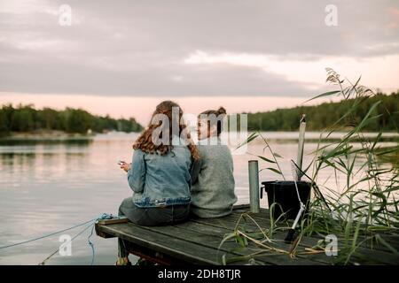 Fille souriante parlant à un frère tout en étant assise sur la jetée pendant coucher de soleil Banque D'Images