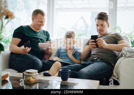 Père souriant avec smartphone regardant son fils utilisant le numérique tablette sur le canapé à la maison Banque D'Images