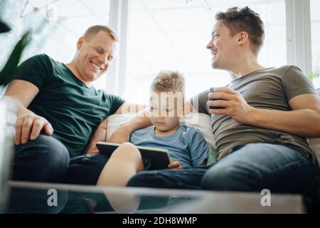 Les pères homosexuels souriants parlent pendant que leur fils utilise une tablette numérique à accueil Banque D'Images