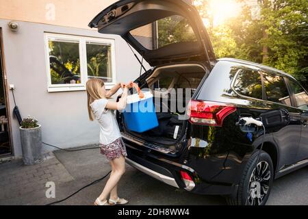 Fille chargement de refroidisseur dans le coffre de voiture noir contre la maison Banque D'Images