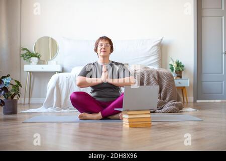 Une femme âgée médite dans lotus poser à la maison devant un moniteur d'ordinateur portable. Le concept d'un mode de vie sain et actif dans la vieillesse. Banque D'Images