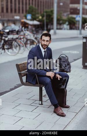 Chef d'entreprise avec sac et lime en regardant loin tout en étant assis sur banc en ville Banque D'Images