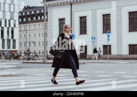 Femme d'affaires qui envisage de traverser la route avec un téléphone intelligent en ville Banque D'Images