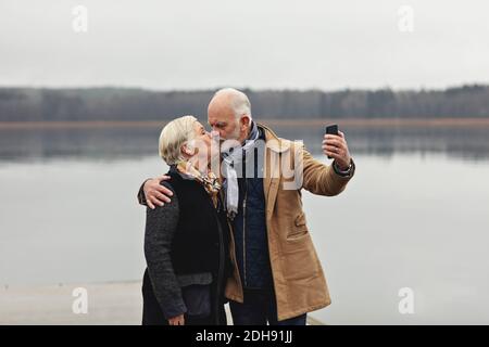 Couple senior embrassant tout en prenant selfie par le lac contre clair ciel Banque D'Images