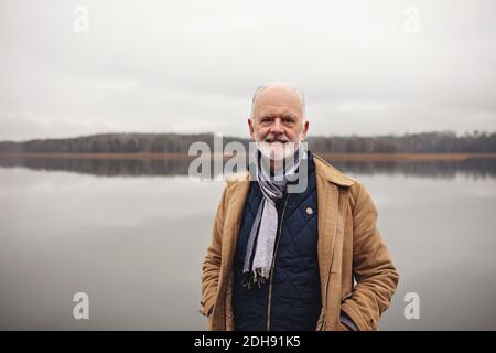 Portrait d'un homme âgé souriant avec les mains dans les poches debout contre le lac Banque D'Images