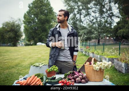 Homme adulte moyen ayant du café tout en se tenant à côté fraîchement moissonné légumes sur table dans le jardin urbain Banque D'Images