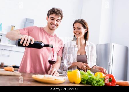 Homme souriant versant du vin près de la petite amie avec un couteau et des légumes sur un premier plan flou Banque D'Images