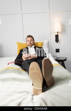 Homme d'affaires se détendant au lit tout en écoutant de la musique depuis son mobile téléphone dans la chambre d'hôtel Banque D'Images