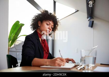 Une jeune femme confiante et confiante en matière de finances écrit dans un journal tout en étant assise avec ordinateur portable au bureau Banque D'Images