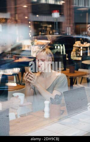 Jeune client androgyne utilisant un smartphone lorsqu'il est assis dans un café vue depuis la fenêtre Banque D'Images