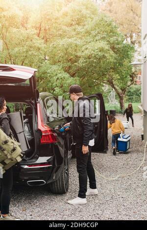 Homme de taille moyenne chargé de la voiture électrique pendant que la famille charge les bagages pendant le pique-nique Banque D'Images