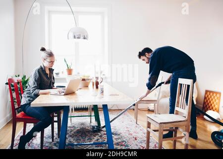 Femme utilisant un ordinateur portable alors qu'un homme nettoie la moquette avec un aspirateur à la maison Banque D'Images