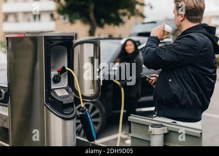 Homme buvant du café en se tenant à la station de charge de voiture électrique Banque D'Images