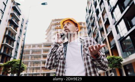 Un architecte mâle en colère dans un casque orange dur parle des oreilles au téléphone sur le chantier de construction. Le créateur en chemise à motif écossais displase émotionnellement Banque D'Images