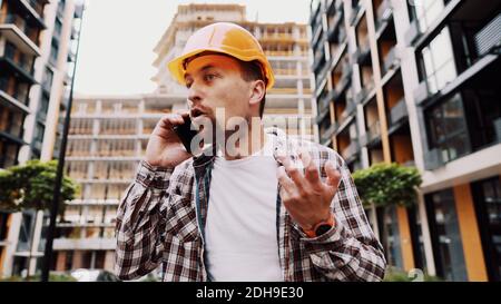 Un architecte mâle en colère dans un casque orange dur parle des oreilles au téléphone sur le chantier de construction. Le créateur en chemise à motif écossais displase émotionnellement Banque D'Images