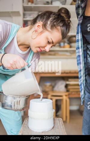 Jeune femelle potter versant l'argile du pichet dans un vase tabouret par une femme debout à l'atelier Banque D'Images