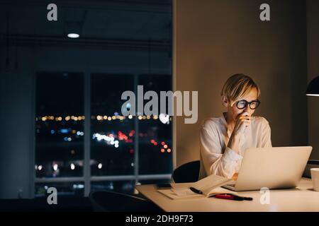 Femme d'affaires mature et dévouée utilisant un ordinateur portable tout en étant assise sur un bureau éclairé dans l'espace de travail des collègues Banque D'Images