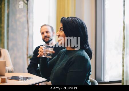 Femme d'affaires souriante tenant un verre d'eau tout en étant assise à la conférence table avec des collègues Banque D'Images