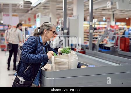 Femme d'âge mûr mettant des produits d'épicerie au comptoir de caisse dans le supermarché Banque D'Images