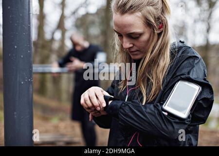 Une athlète féminine vérifie une montre intelligente dans la forêt alors qu'un homme se tient debout en arrière-plan Banque D'Images