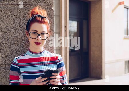 Portrait d'une jeune fille hipster tenant un téléphone portable debout contre le mur Banque D'Images