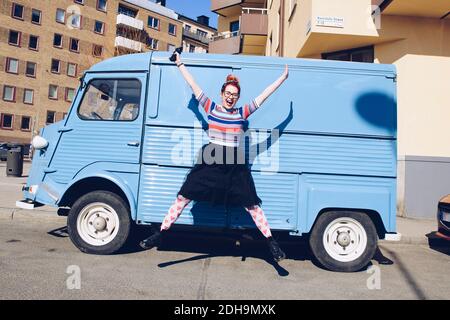 Pleine longueur de jeune femme sautant contre un mini-van bleu sur la rue de la ville Banque D'Images