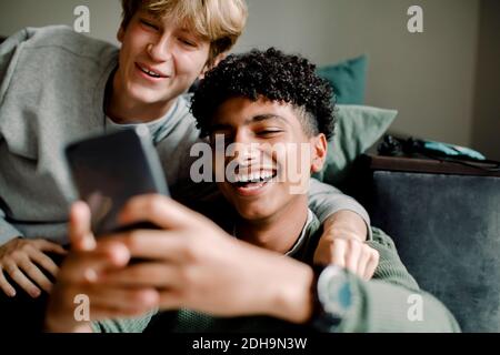 Messagerie texte d'un jeune garçon souriant via un smartphone en étant assis avec un ami à la maison Banque D'Images