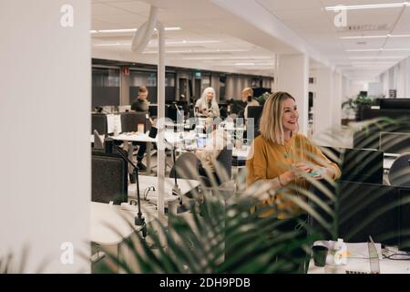 Femme d'affaires souriante qui regarde loin en se tenant au bureau Banque D'Images
