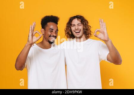 Joyeux multiculturel deux gars montrant le signe ok et wencant pendant embrassant isolé sur fond jaune Banque D'Images