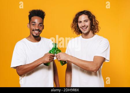 Heureux multiculturel deux gars souriant et clinking bouteilles isolées arrière-plan jaune Banque D'Images