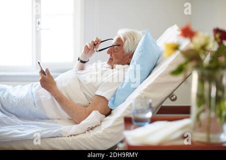 Vue latérale d'un homme âgé portant des lunettes de vue tout en utilisant Smart téléphone dans le service hospitalier Banque D'Images