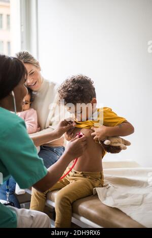 Femme médecin examinant le battement de coeur du garçon avec stéthoscope pendant que la mère est assise et fille en clinique Banque D'Images