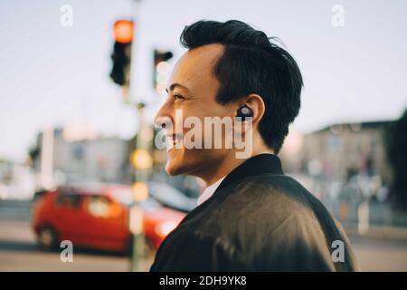 Homme plein de gaieté qui regarde loin en portant des écouteurs intra-auriculaires en ville Banque D'Images